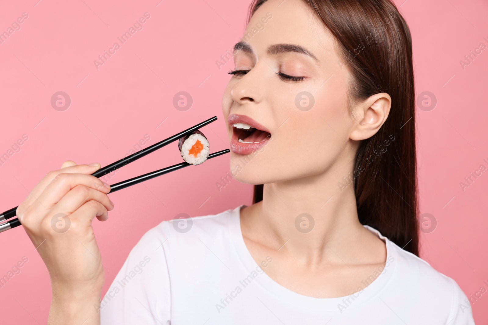 Photo of Beautiful young woman eating sushi roll with chopsticks on pink background