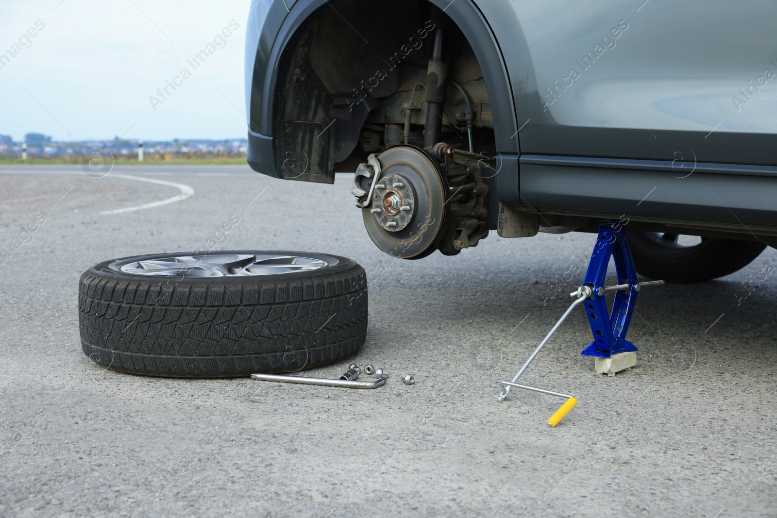 Photo of Car lifted by scissor jack and new wheel on roadside outdoors. Tire puncture