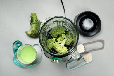 Making shake. Broccoli in blender and scoops with different powders on light gray table, flat lay