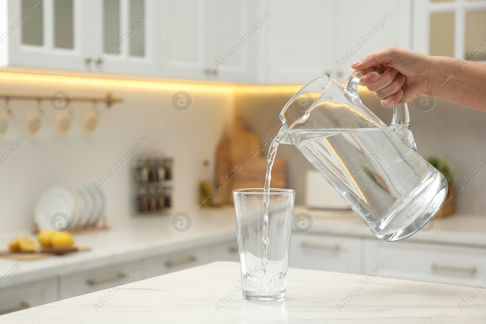 Photo of Woman pouring water from jug into glass at white table in kitchen, closeup. Space for text