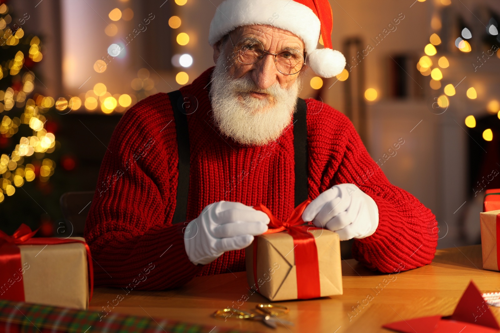 Photo of Santa Claus tying bow on gift box at his workplace in room decorated for Christmas