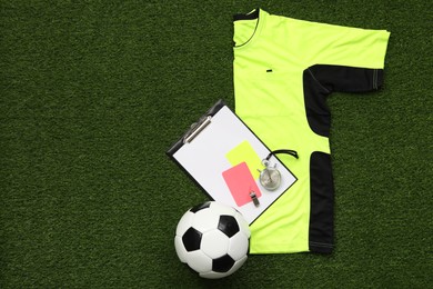 Uniform, soccer ball and other referee equipment on green grass, top view. Space for text