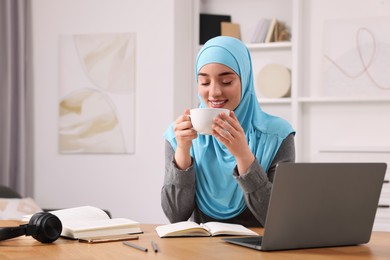 Photo of Muslim woman in hijab with cup of coffee using laptop at wooden table in room