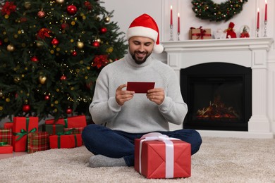 Happy young man in Santa hat with gift boxes reading greeting card in room decorated for Christmas