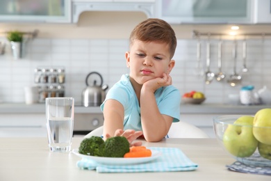 Adorable little boy refusing to eat vegetables at table in kitchen