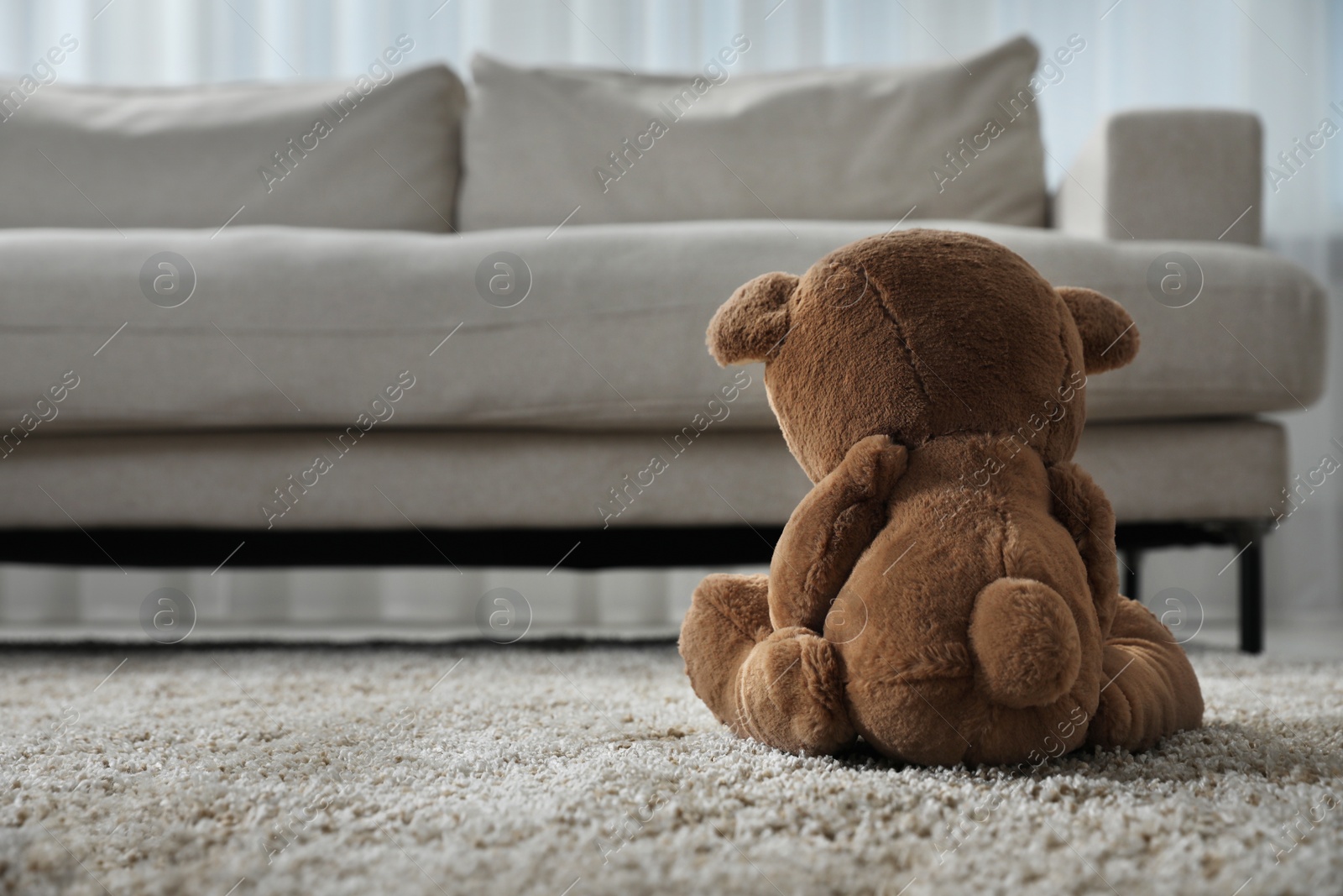 Photo of Cute lonely teddy bear on floor near sofa in room, back view. Space for text
