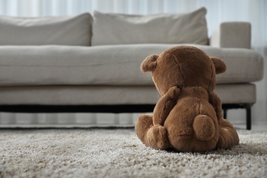 Cute lonely teddy bear on floor near sofa in room, back view. Space for text