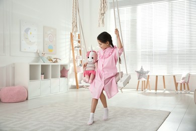 Cute little girl playing with toy unicorn on swing at home