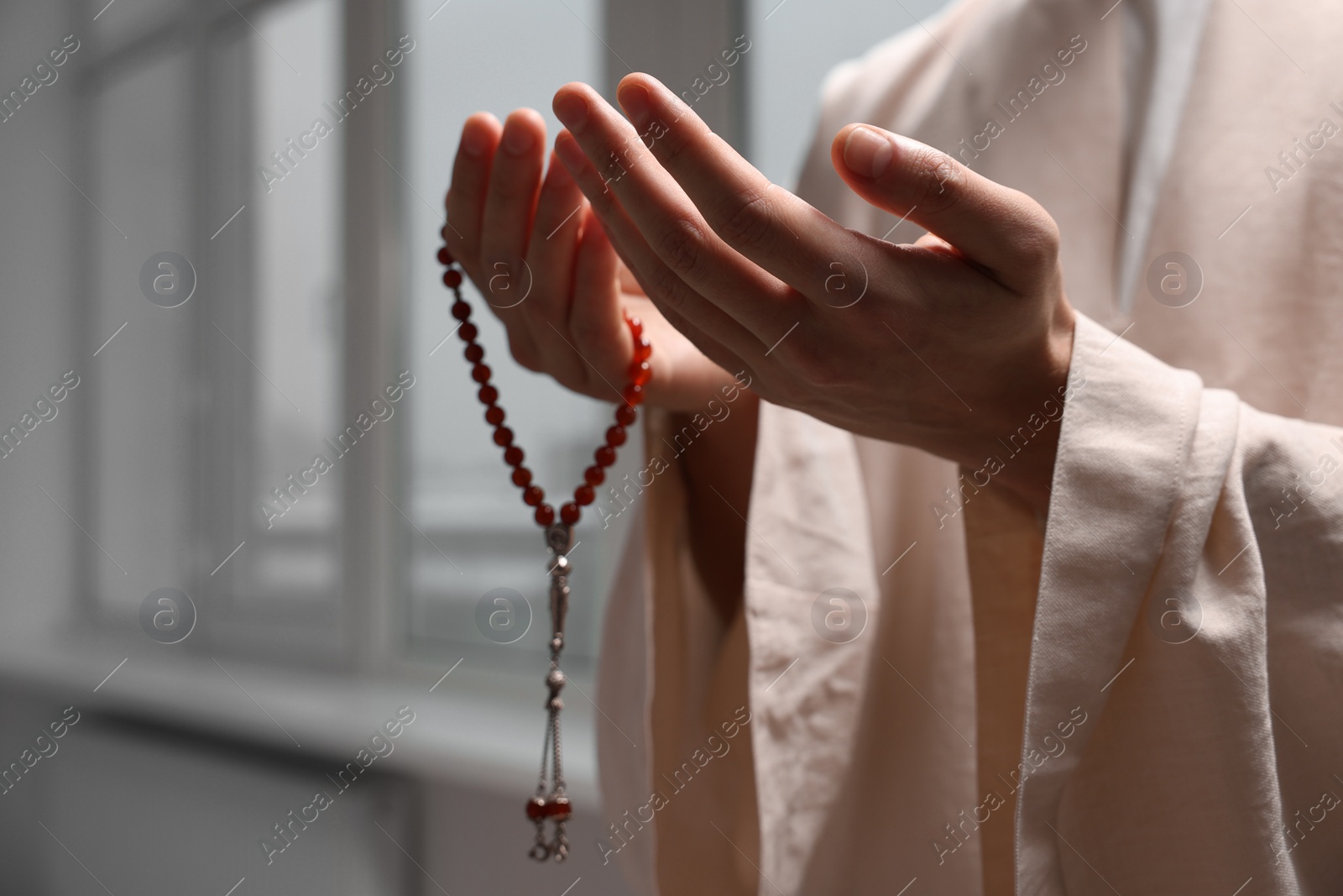 Photo of Muslim man with misbaha praying near window indoors, closeup. Space for text