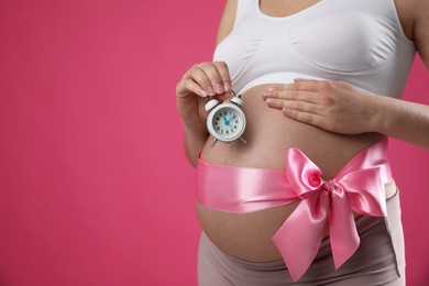 Photo of Young pregnant woman with alarm clock and bow on pink background, closeup. Time to give birth