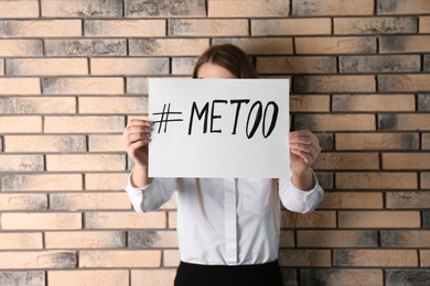 Woman holding paper with text "#METOO" near brick wall. Problem of sexual harassment at work