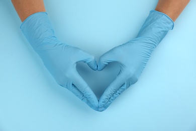 Person in medical gloves making heart with hands on light blue background, top view