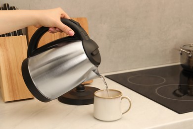 Photo of Woman pouring hot water from electric kettle into cup in kitchen, closeup