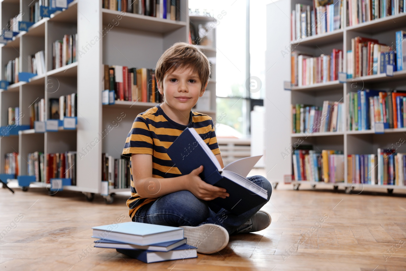 Photo of Cute little boy reading book on floor in library