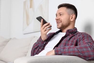Photo of Handsome man recording voice message via smartphone on sofa at home. Space for text