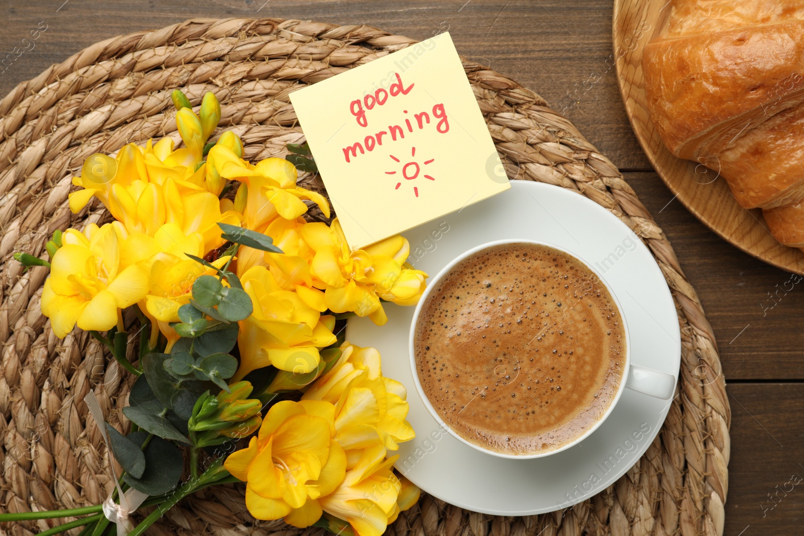 Photo of Cup of aromatic coffee with croissant, beautiful yellow freesias and Good Morning note on wooden table, flat lay