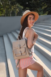Photo of Beautiful young woman with stylish backpack and hat on stairs outdoors