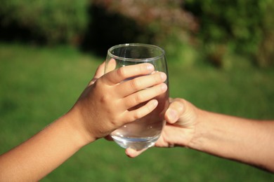 Photo of Child giving glass of water to elderly woman outdoors on sunny day, closeup
