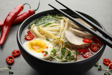 Delicious ramen with meat in bowl, chopsticks and chili peppers on grey textured table. Noodle soup