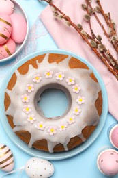 Delicious Easter cake decorated with sprinkles near painted eggs and willow branches on light blue background, flat lay