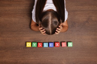 Photo of Little girl covering head with hands and cubes with word Dyslexia at wooden table, top view