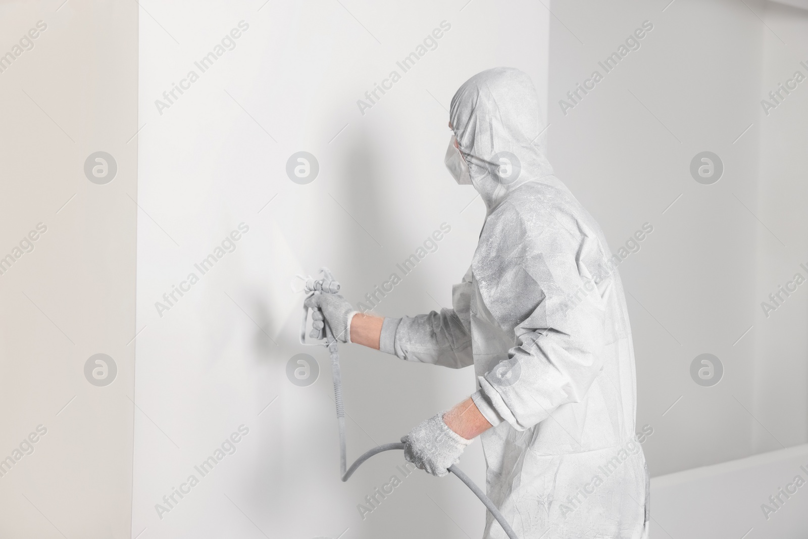 Photo of Decorator in protective overalls painting wall with spray gun indoors