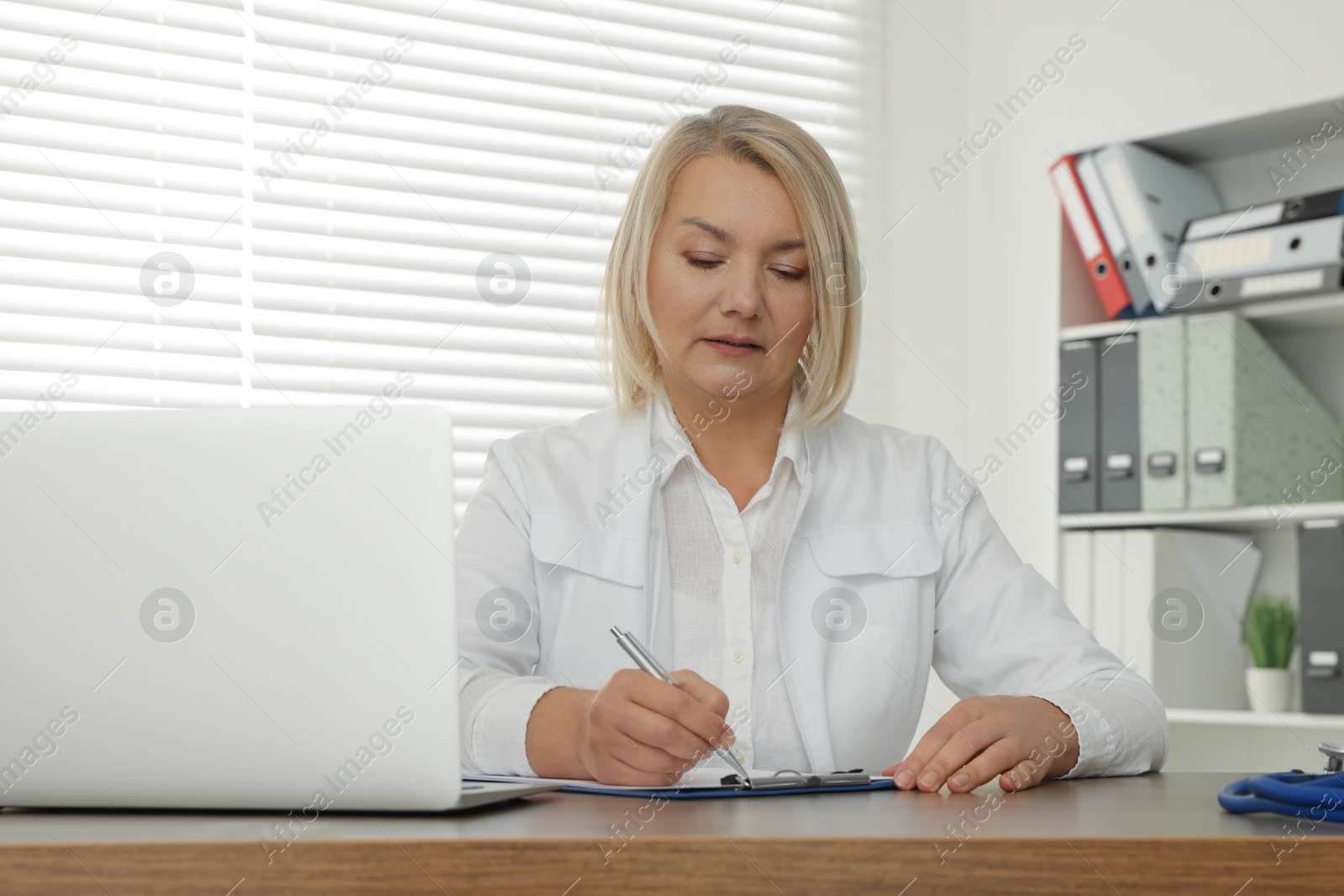 Photo of Doctor with laptop and clipboard writing at wooden table in clinic