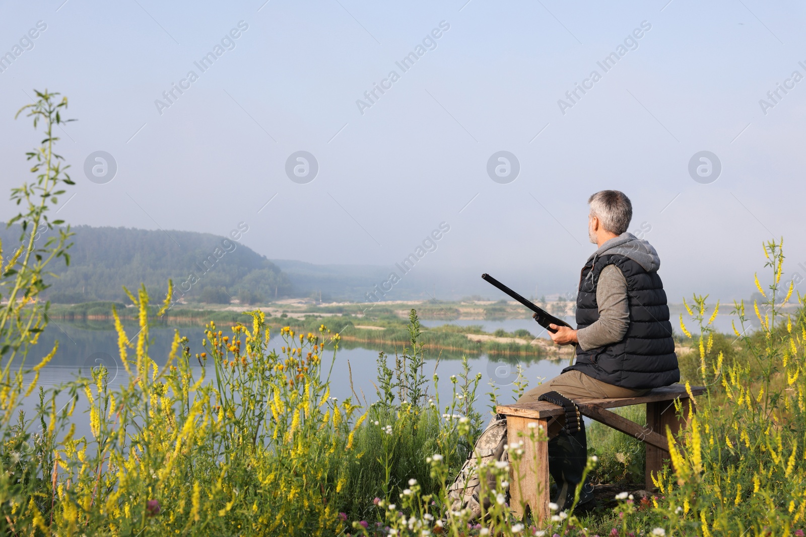 Photo of Man with hunting rifle sitting on wooden bench near lake outdoors. Space for text