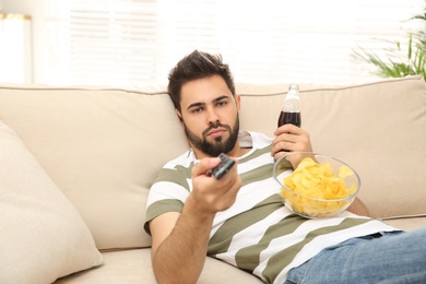 Lazy young man with chips and drink watching TV on sofa at home