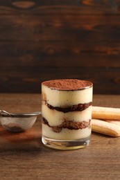 Photo of Delicious tiramisu in glass, biscuits and sieve with cocoa powder on wooden table