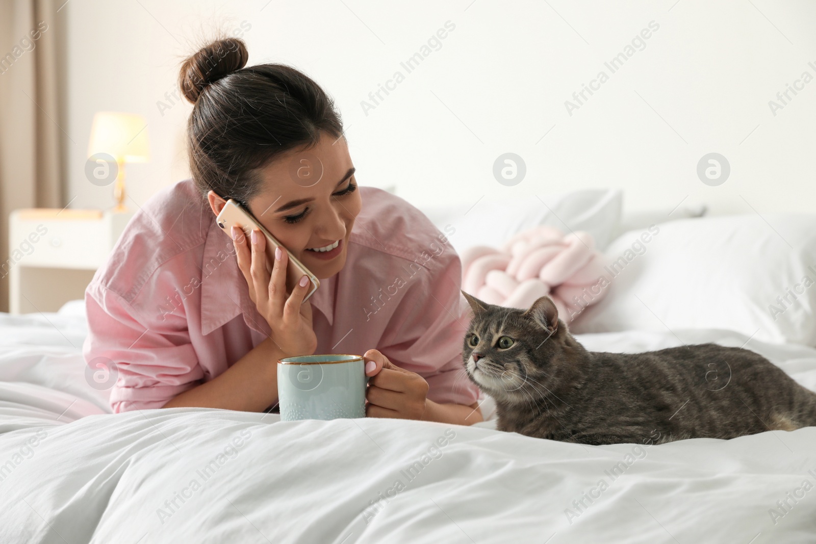 Photo of Young woman with cup of coffee talking on phone while lying near cute cat in bedroom. Pet and owner