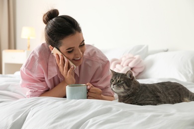 Young woman with cup of coffee talking on phone while lying near cute cat in bedroom. Pet and owner