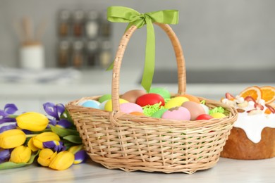 Photo of Easter basket with painted eggs between tulips and cake on white marble table indoors