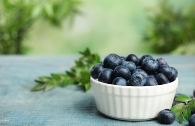 Bowl of tasty blueberries and leaves on wooden table against blurred green background, space for text