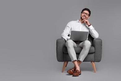 Happy man with laptop sitting in armchair on grey background. Space for text
