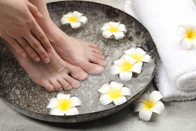 Photo of Woman soaking her feet in bowl with water and flowers on light grey floor, closeup. Spa treatment