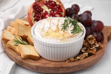Photo of Board with tasty baked camembert, croutons, grapes, walnuts and pomegranate on white tiled table, closeup