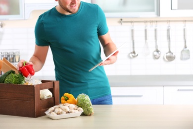 Photo of Man with wooden crate full of products and tablet in kitchen, space for text. Food delivery service