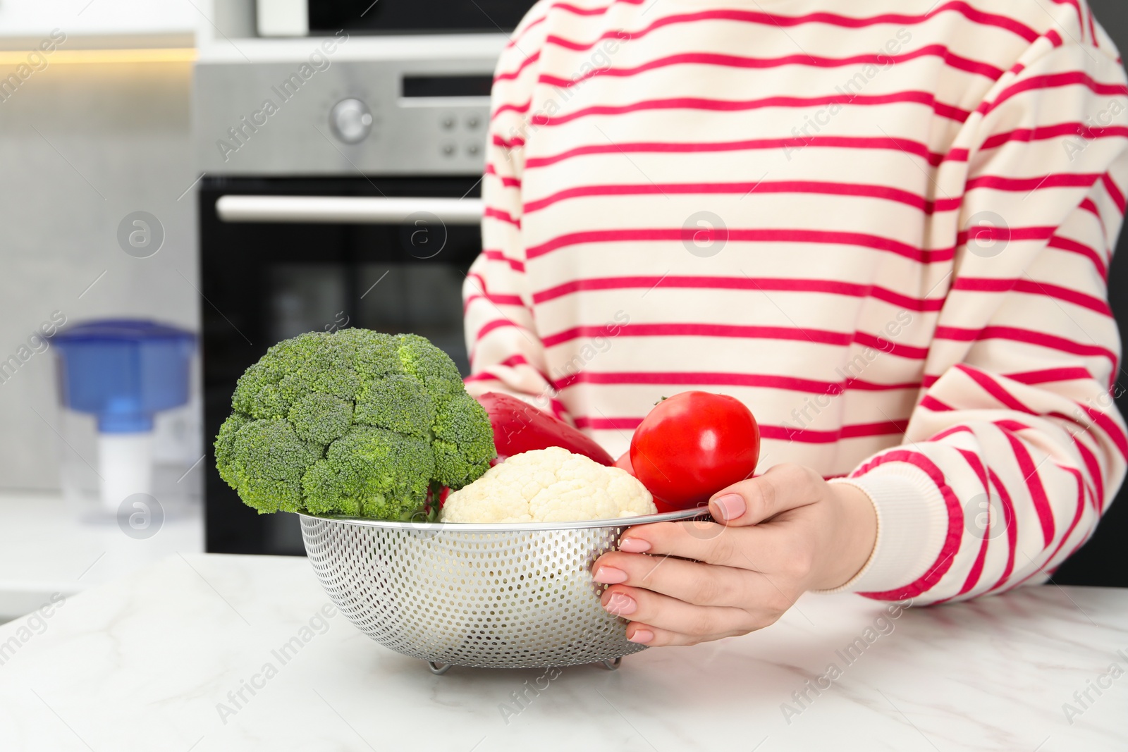 Photo of Woman holding colander with fresh vegetables at white marble table in kitchen, closeup