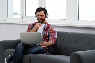 Smiling bearded man with headphones and laptop on sofa indoors