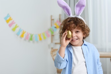 Photo of Happy boy in cute bunny ears headband covering eye with Easter egg indoors. Space for text