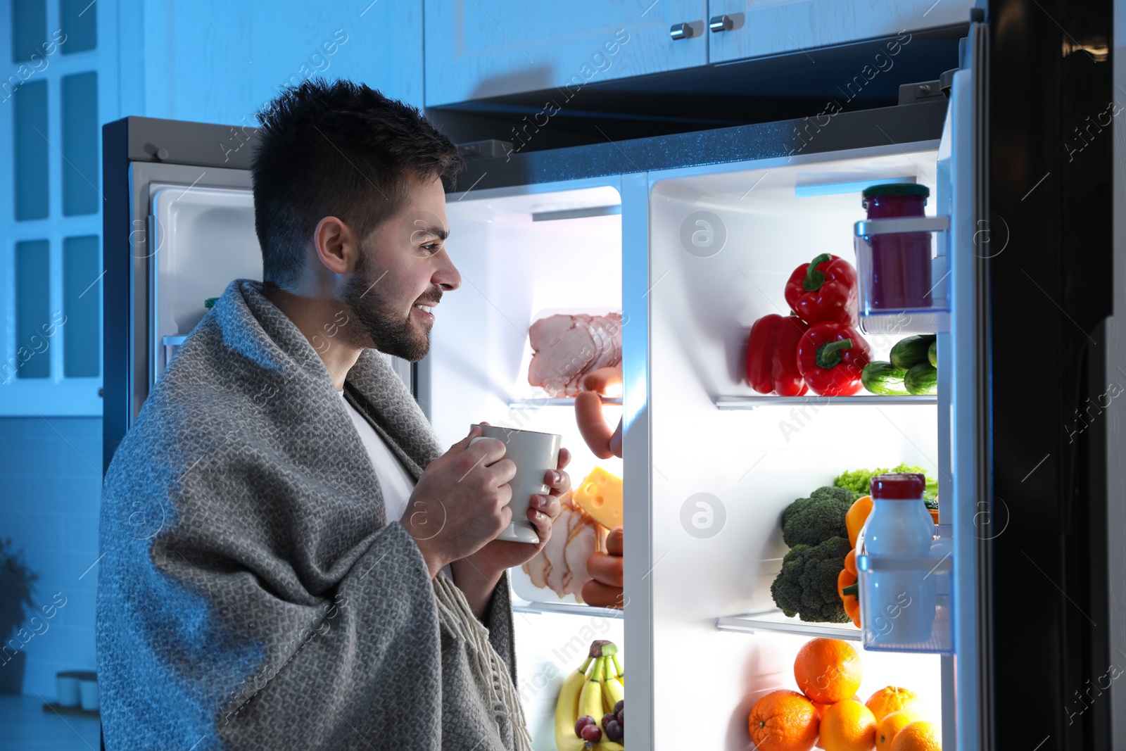 Photo of Young man with cup of drink near open refrigerator at night
