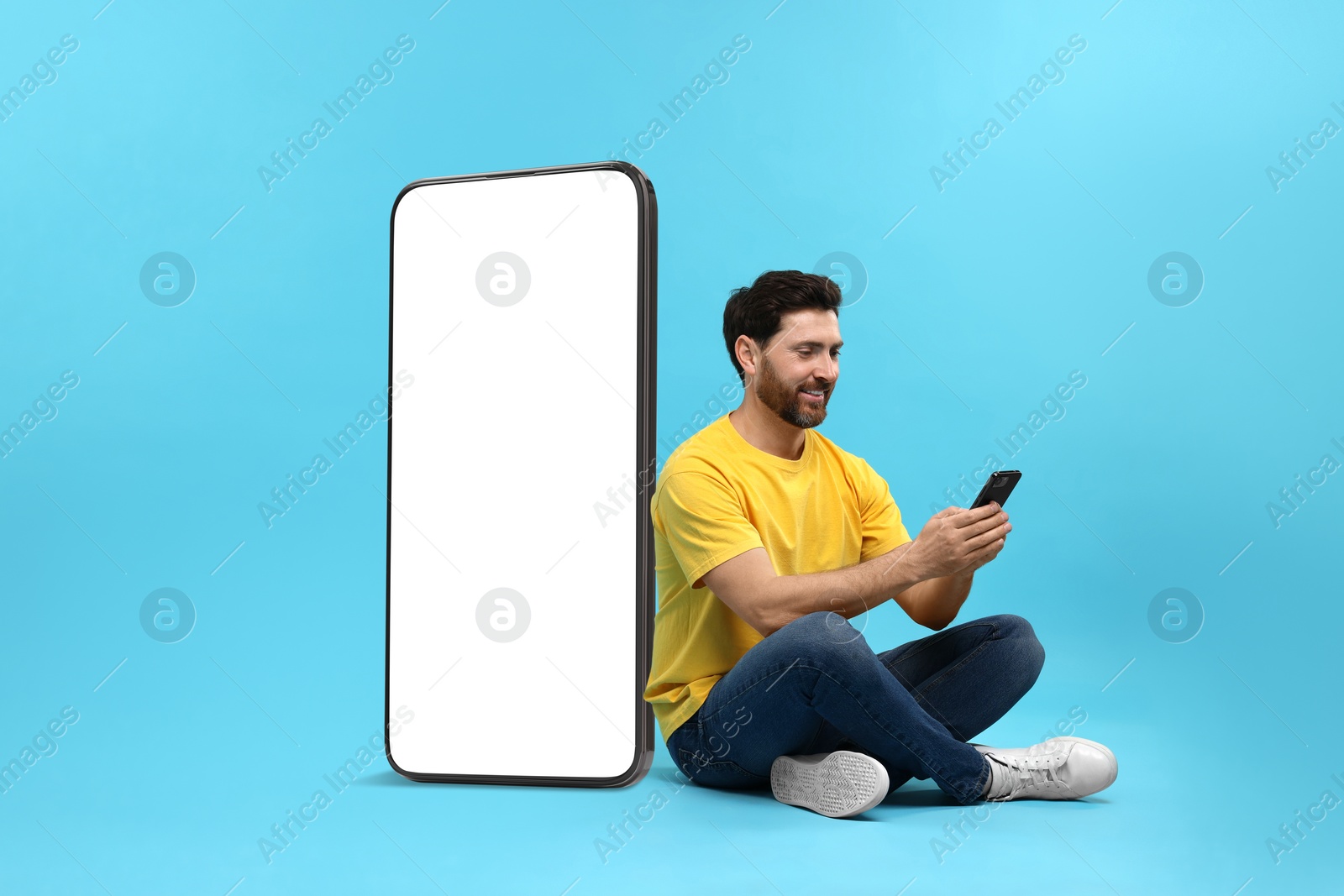 Image of Man with mobile phone sitting near huge device with empty screen on light blue background. Mockup for design