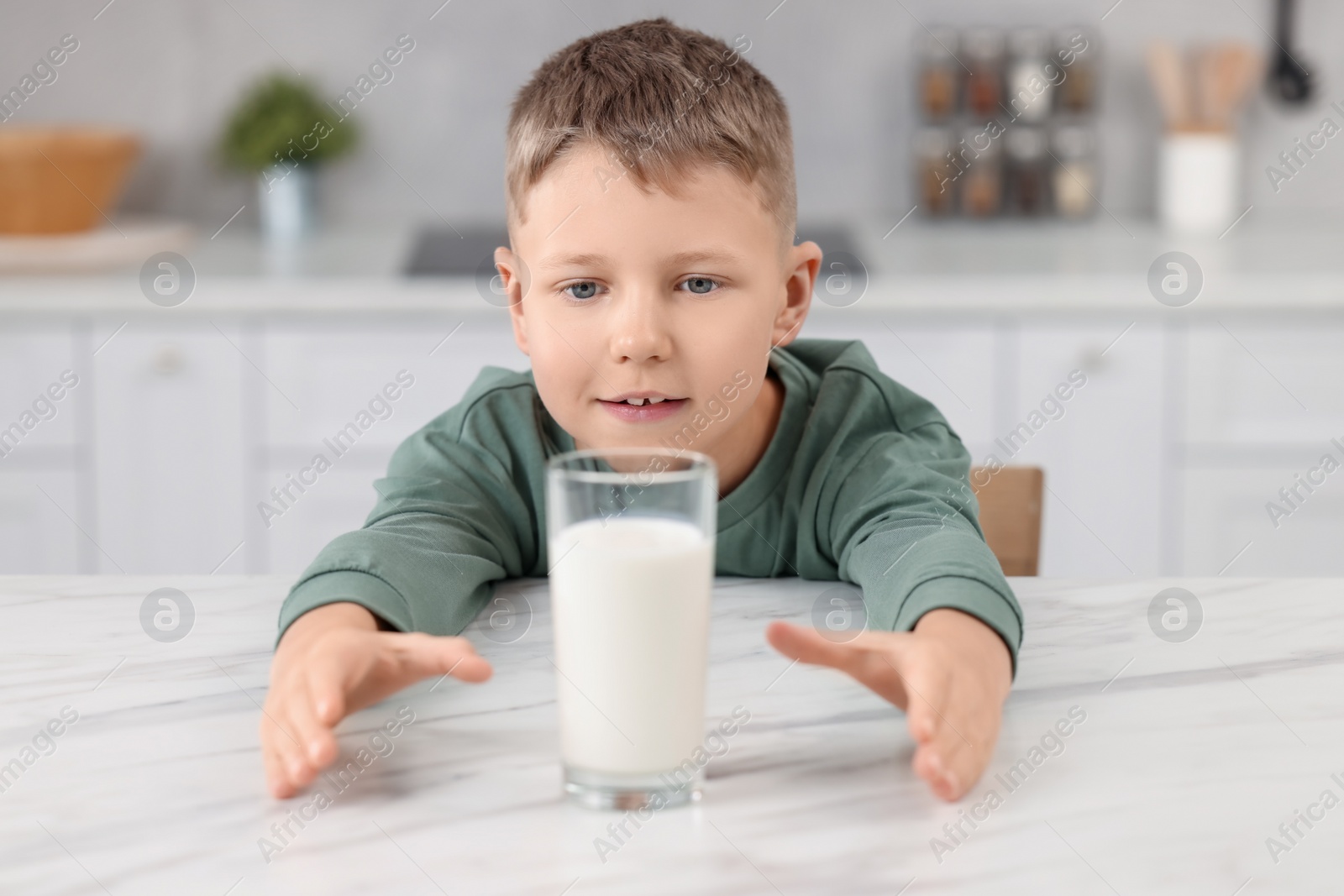 Photo of Cute boy reaching out for glass of milk at white table in kitchen