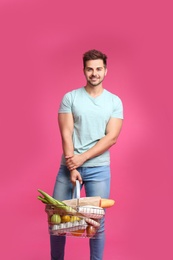 Young man with shopping basket full of products on pink background