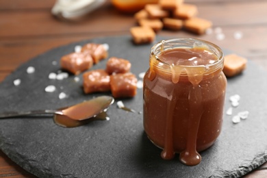 Photo of Slate plate with jar of tasty caramel sauce on wooden table. Space for text
