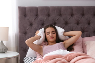 Photo of Young woman covering ears with pillow while trying to sleep in bed at home. Early morning