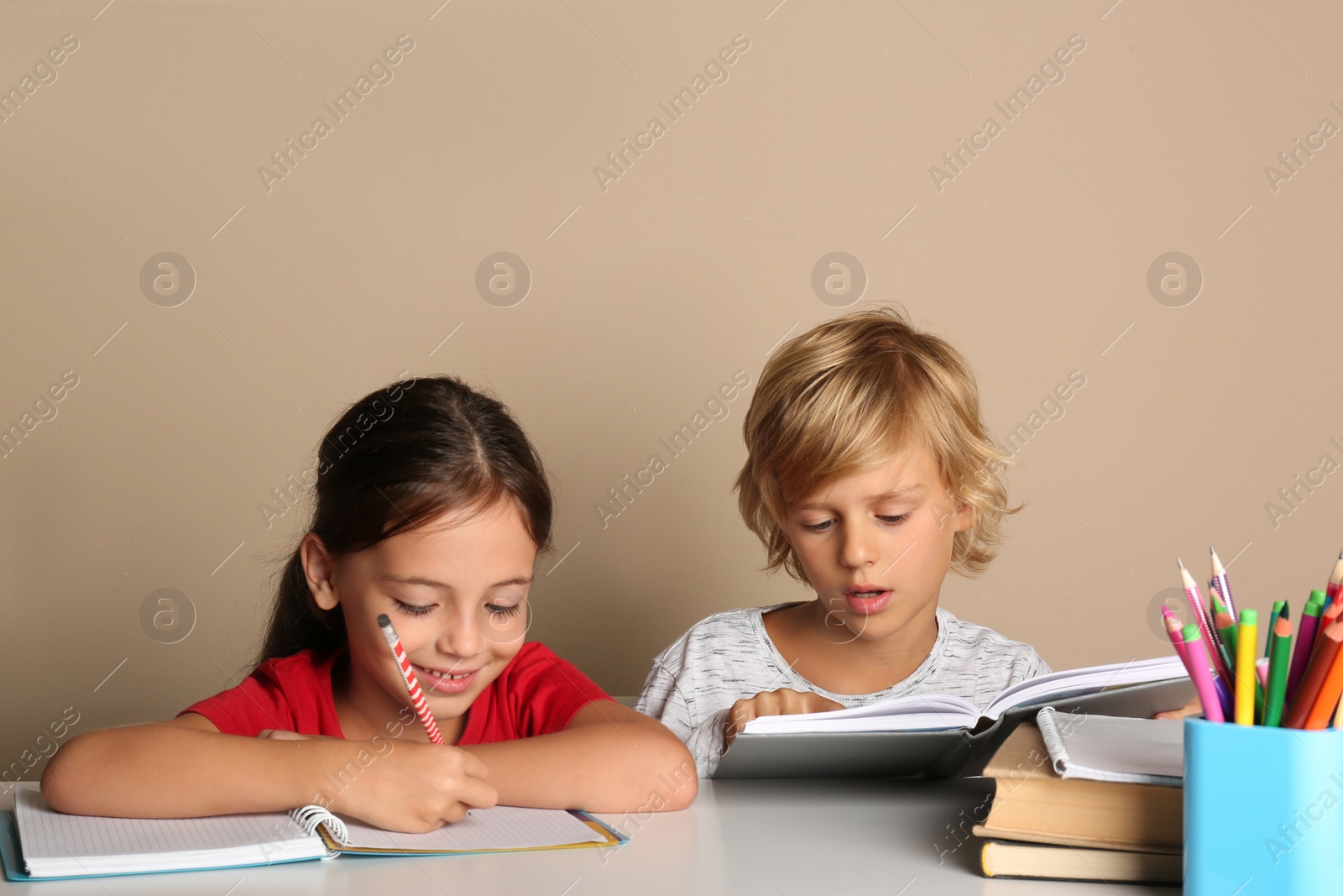 Photo of Little boy and girl doing homework at table on beige background