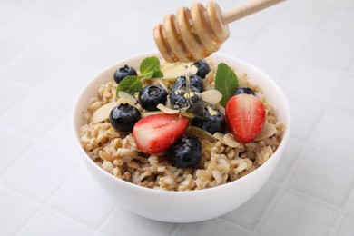 Photo of Pouring honey onto tasty oatmeal with strawberries, blueberries and almond petals in bowl on white tiled table, closeup