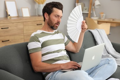 Photo of Bearded man with laptop waving white hand fan to cool himself on sofa at home